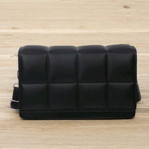 以下が含まれることがあります： キルティングデザインの黒い長方形のクロスボディバッグ。バッグは、前面にパッド入りの正方形のパターンと、細い調節可能なストラップが特徴です。バッグは滑らかでマットな素材で作られています。