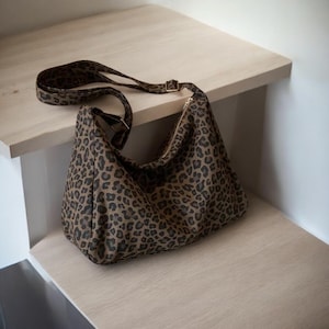 May include: A brown and black leopard print shoulder bag with a long adjustable strap. The bag has a zipper closure and a gold-colored buckle. The bag is sitting on a light-colored wooden surface.