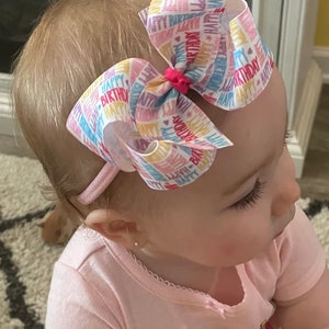 May include: A white headband with a large pink, blue, and yellow bow. The bow is made of ribbon with the words "Happy Birthday" printed on it. The headband is worn by a young child.
