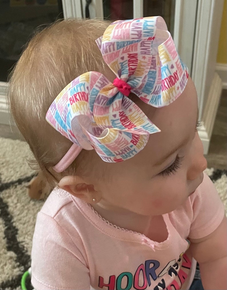 May include: A white headband with a large pink, blue, and yellow bow. The bow is made of ribbon with the words "Happy Birthday" printed on it. The headband is worn by a young child.