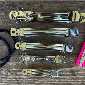 May include: A set of five silver hair clips and a pink hair clip with a black elastic band. The hair clips are different sizes and have a smooth, shiny finish.