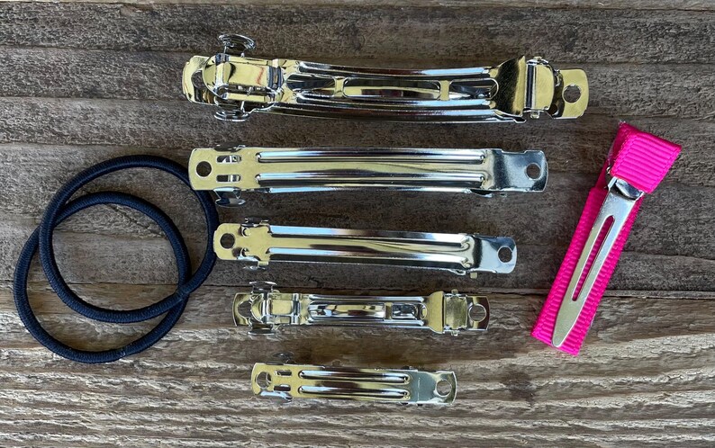 May include: A set of five silver hair clips and a pink hair clip with a black elastic band. The hair clips are different sizes and have a smooth, shiny finish.