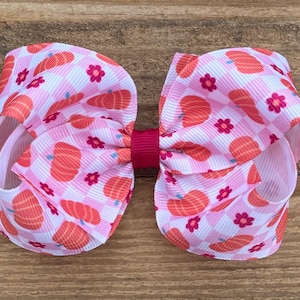 May include: A large pink and white hair bow with a pumpkin and gingham pattern. The bow is tied with a bright pink ribbon.