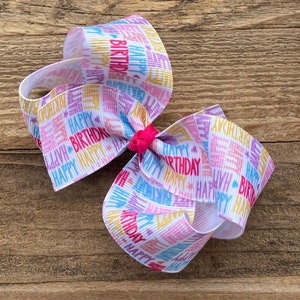 May include: A white hair bow with a colorful "Happy Birthday" print. The bow is made of ribbon and has a pink center.