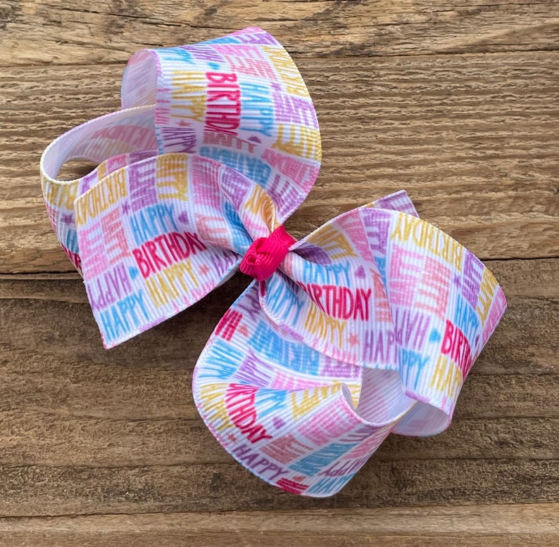 May include: A white hair bow with a colorful "Happy Birthday" print. The bow is made of ribbon and has a pink center.