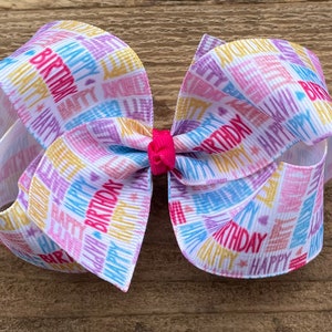 May include: A white hair bow with a colorful "Happy Birthday" print. The bow is tied with a pink ribbon.