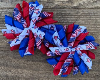 May include: Two red, white, and blue ribbon hair bows with a star pattern. The bows are curled and tied together.