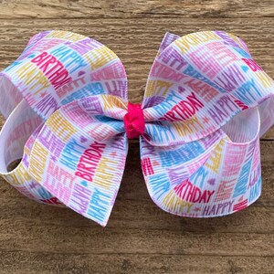 May include: A white hair bow with a colorful "Happy Birthday" print. The bow is tied with a pink ribbon in the center.