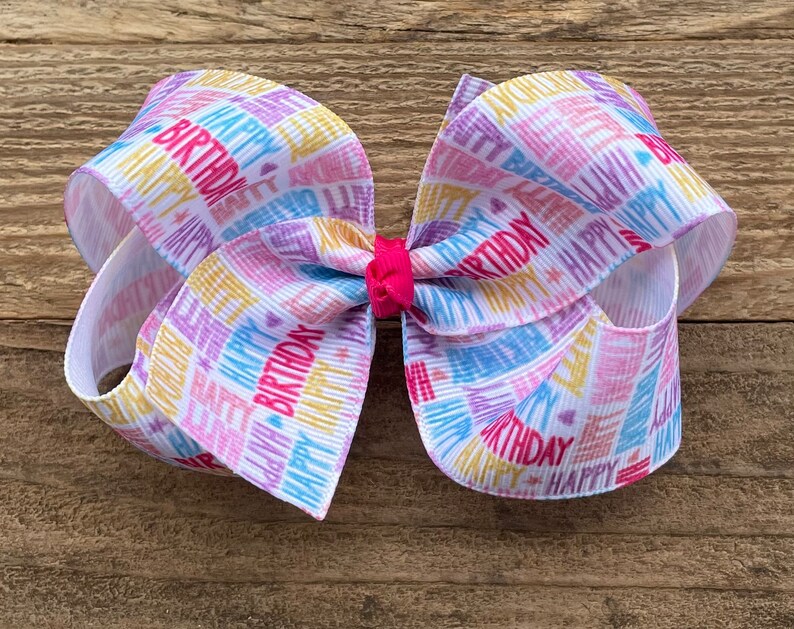 May include: A white hair bow with a colorful "Happy Birthday" print. The bow is tied with a pink ribbon in the center.