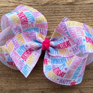 May include: A white hair bow with a colorful "Happy Birthday" print. The bow is made of ribbon and has a pink center.