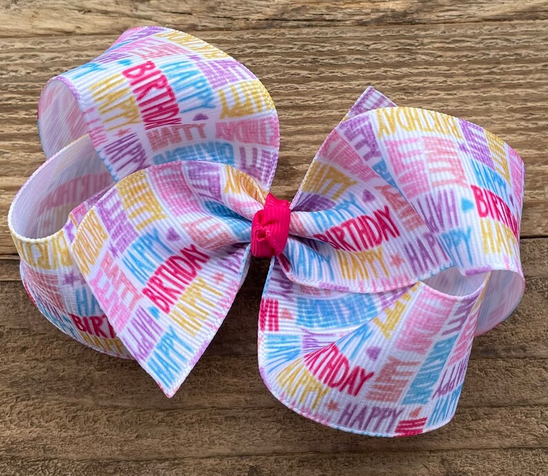 May include: A white hair bow with a colorful "Happy Birthday" print. The bow is made of ribbon and has a pink center.
