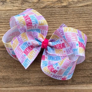 May include: A white hair bow with a colorful "Happy Birthday" print. The bow is made of ribbon and has a pink center.