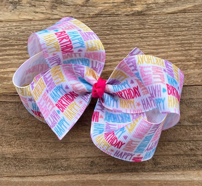 May include: A white hair bow with a colorful "Happy Birthday" print. The bow is made of ribbon and has a pink center.