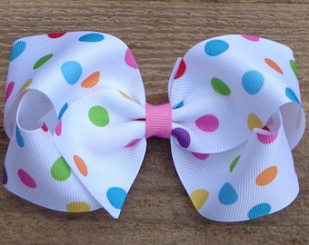 May include: A white hair bow with a polka dot pattern in various colors, including red, orange, yellow, green, blue, and purple. The bow is tied with a pink ribbon in the center.