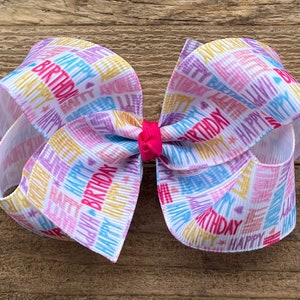 May include: A white, pink, blue, and yellow birthday ribbon bow with the words "Happy Birthday" printed on it.