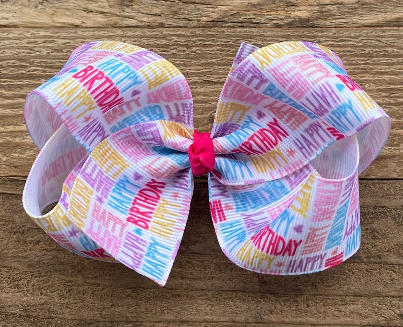 May include: A white, pink, blue, and yellow birthday ribbon bow with the words "Happy Birthday" printed on it.