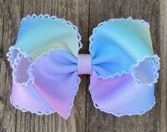 May include: A pastel rainbow ombre hair bow with white lace trim. The bow is made of a soft, textured fabric and has a center loop for attaching to hair.