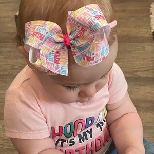 May include: A baby girl wearing a pink shirt with the text "HOOP IT'S MY 1ST BTHDA" in colorful letters. She is wearing a headband with a colorful bow that says "Happy Birthday" all over it. She is playing with a black toy.