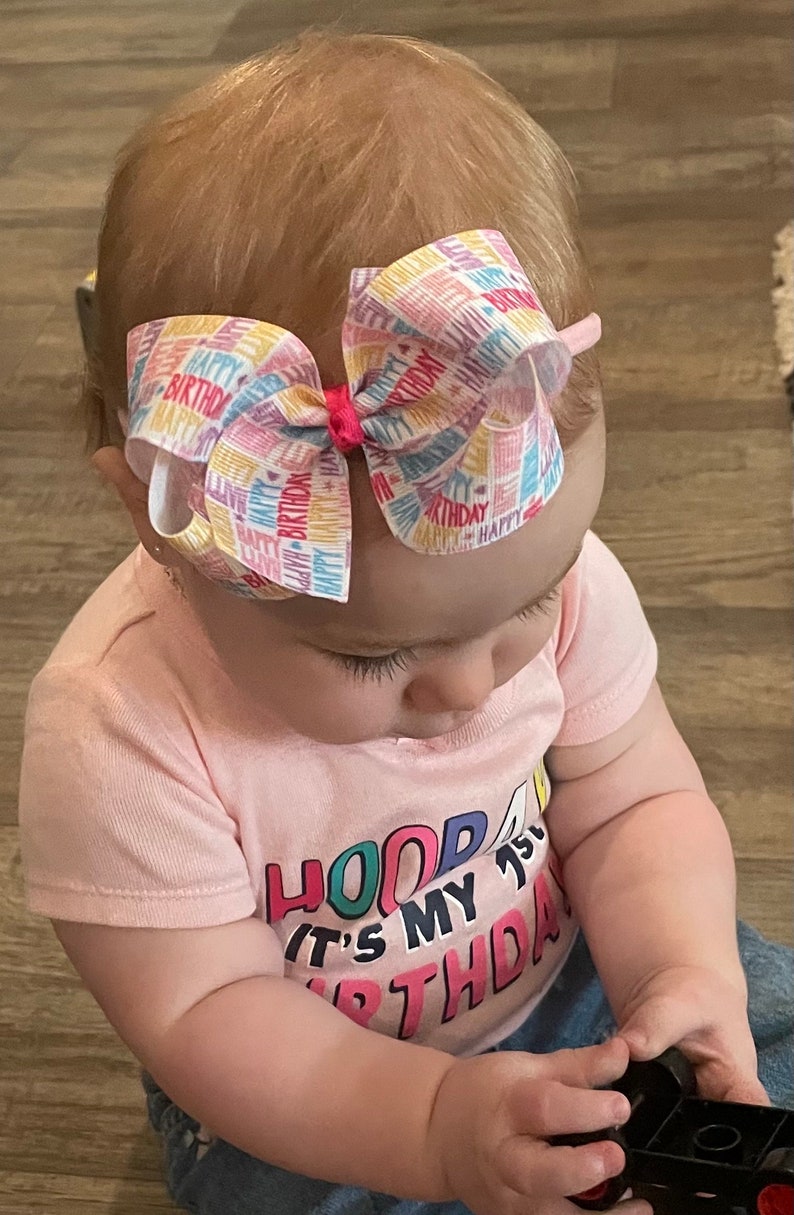 May include: A baby girl wearing a pink shirt with the text "HOOP IT'S MY 1ST BTHDA" in colorful letters. She is wearing a headband with a colorful bow that says "Happy Birthday" all over it. She is playing with a black toy.