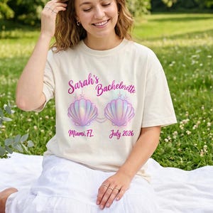 以下が含まれることがあります： アイボリー色のTシャツ。ピンクと紫の貝殻のグラフィックと「Sarah's Bachelorette」の文字がプリントされています。「Miami, FL July 2026」の文字もプリントされています。芝生の上で白いブランケットに座っている人。