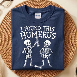 May include: A navy blue crew neck t-shirt featuring a white graphic of two skeletons and the text I FOUND THIS HUMERUS. The design shows one skeleton holding a bone up while the other stands with hands on hips, with another bone on the ground.