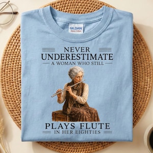 Peut inclure: Un t-shirt bleu clair avec une illustration d'une personne âgée jouant de la flûte. Le texte indique : NEVER UNDERESTIMATE A WOMAN WHO STILL PLAYS FLUTE IN HER EIGHTIES. Le vêtement présente un col rond et une étiquette Gildan à l'intérieur.