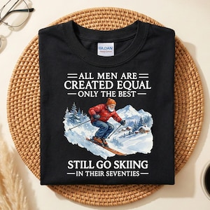 Puede incluir: Camiseta negra con el texto "ALL MEN ARE CREATED EQUAL - ONLY THE BEST - STILL GO SKIING - IN THEIR SEVENTIES." La camiseta presenta un gráfico de un esquiador con chaqueta roja y pantalones azules. La camiseta está sobre una estera tejida.