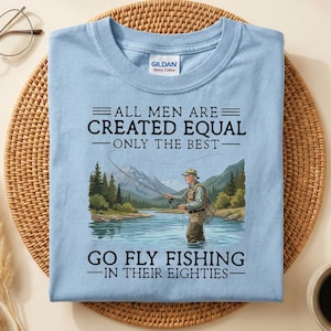 May include: Light blue t-shirt with the text "ALL MEN ARE CREATED EQUAL - ONLY THE BEST - GO FLY FISHING - IN THEIR EIGHTIES." The graphic shows a man fly fishing in a river with mountains in the background.