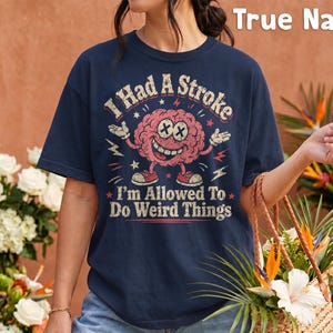 Puede incluir: Una camiseta azul marino con un gráfico de un cerebro rosa con ojos en forma de X y una amplia sonrisa. El texto dice I Had A Stroke I'm Allowed To Do Weird Things. El diseño incluye pequeñas estrellas y rayos decorativos.