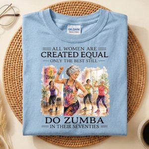 May include: A light blue t-shirt featuring a graphic print of three individuals dancing. The text reads: ALL WOMEN ARE CREATED EQUAL ONLY THE BEST STILL DO ZUMBA IN THEIR SEVENTIES. The shirt has a Gildan brand label at the inner neckline.