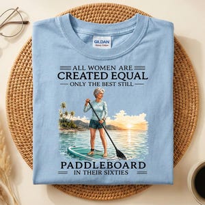 May include: A light blue t-shirt featuring a graphic of a person on a paddleboard at sunset. The text reads: ALL WOMEN ARE CREATED EQUAL ONLY THE BEST STILL PADDLEBOARD IN THEIR SIXTIES. The shirt has a Gildan brand label at the inner neckline.