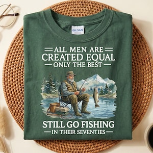 Op de afbeelding: Een donkergroen t-shirt met de tekst "ALL MEN ARE CREATED EQUAL - ONLY THE BEST - STILL GO FISHING - IN THEIR SEVENTIES." De afbeelding toont een oudere man die aan het vissen is. Het shirt is op een geweven mat gevouwen.