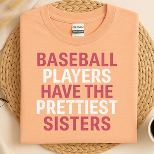 Op de afbeelding: Perzikkleurig T-shirt met de tekst "BASEBALL PLAYERS HAVE THE PRETTIEST SISTERS" in wit en bordeauxrood. Het shirt is opgevouwen op een geweven mat, met een kopje koffie en een bril in de buurt.