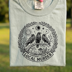 以下が含まれることがあります： 丸首の薄緑色のTシャツ。黒と白のグラフィックで、3羽のカラスが枝に止まっている様子が描かれています。太陽、月、星が周りに描かれ、下には「Support Your Local Murder」の文字がプリントされています。