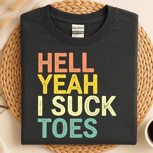 Może przedstawiać: Czarny t-shirt z napisem "HELL YEAH I SUCK TOES" w kolorowych blokowych literach. Koszulka wykonana jest z ciężkiej bawełny i ma okrągły dekolt. Koszulka jest prezentowana na tkanej macie.