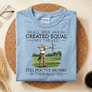 May include: Light blue t-shirt with the text "ALL MEN ARE CREATED EQUAL - ONLY THE BEST - STILL PRACTICE ARCHERY - IN THEIR EIGHTIES." The graphic shows an archer aiming at a target. The shirt is made of a soft, comfortable material.