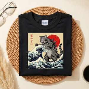 Può includere: T-shirt nero con una grafica di un gatto simile a Godzilla, ambientato contro un sole rosso e onde oceaniche. Il design include testo giapponese. La maglietta è realizzata in un materiale morbido.