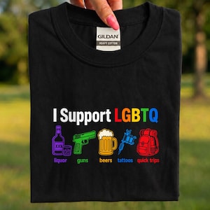 May include: Black t-shirt with the text "I Support LGBTQ" in white and rainbow colors. Below the text are colorful illustrations of liquor, a gun, beer, tattoos, and a backpack. The shirt is made of heavy cotton.