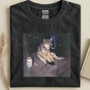 May include: Dark gray t-shirt featuring a graphic of a wolf smoking a cigarette, with a beer can and a cigarette on the ground. The wolf is sitting down with a relaxed posture. The shirt is made of heavy cotton.