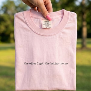 Peut inclure: Un t-shirt à manches courtes rose clair avec une encolure ronde. Le t-shirt présente le texte imprimé en noir "the older I get, the heller the no" centré sur la poitrine. Une étiquette de marque blanche est fixée à l'intérieur du col.