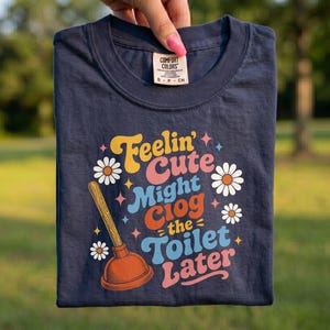 Op de afbeelding: Een donkerblauw t-shirt met korte mouwen en een grafische print met de tekst Feelin' Cute Might Clog the Toilet Later. Het ontwerp bevat een illustratie van een ontstopper, witte madeliefjes en kleine sterretjes in geel, roze en lichtblauw.