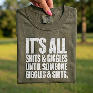 Op de afbeelding: Een saliegroen T-shirt met korte mouwen en witte, verweerde tekst die luidt: IT'S ALL SHITS & GIGGLES UNTIL SOMEONE GIGGLES & SHITS. Het T-shirt heeft een ronde hals en een Comfort Colors-merklabel zichtbaar aan de binnenkant van de kraag.
