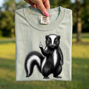 Puede incluir: Una camiseta de color verde salvia claro con una ilustración gráfica en blanco y negro de una mofeta de pie sobre sus patas traseras levantando el dedo corazón. La prenda tiene cuello redondo y una etiqueta de Comfort Colors en el cuello.