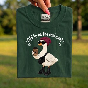 May include: A dark green t-shirt featuring a graphic of a white goose wearing a maroon beanie, black sunglasses, a black fanny pack, and black boots while holding a coffee cup. The text reads, Off to be the cool aunt, with small white hearts.