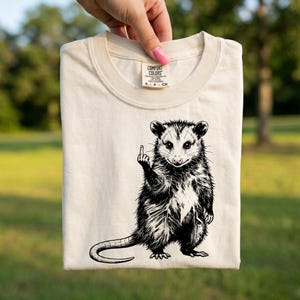 Peut inclure: T-shirt crème avec une illustration en noir et blanc d'un opossum faisant un doigt d'honneur. Le t-shirt est en coton Comfort Colors et a un col rond. Le mot "White" est dans le coin supérieur droit.