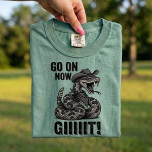 Può includere: Una t-shirt verde chiaro con la grafica di un serpente a sonagli che indossa un cappello da cowboy e una bandana. Il testo sopra il serpente recita GO ON NOW e il testo sotto recita GIIIIT!. La maglietta presenta un'etichetta Comfort Colors.