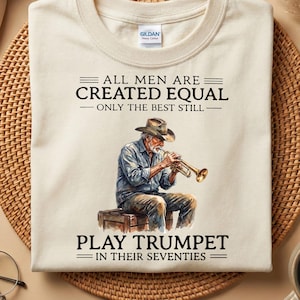 Può includere: T-shirt color crema con grafica di un uomo che suona la tromba, con la scritta "ALL MEN ARE CREATED EQUAL - ONLY THE BEST STILL - PLAY TRUMPET IN THEIR SEVENTIES." La maglietta ha uno scollo tondo.