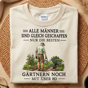 May include: A cream-coloured t-shirt featuring a graphic of a gardener in a vegetable patch with a wheelbarrow and watering can. The text reads: ALLE MÄNNER SIND GLEICH GESCHAFFEN NUR DIE BESTEN GÄRTNERN NOCH MIT ÜBER 80. The shirt has a crew neck.