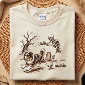 May include: A cream-coloured crew neck t-shirt featuring a graphic print of a skeleton reaching towards a running Australian Shepherd dog that is holding a bone in its mouth. The scene includes a bare tree and a tombstone on a light background.