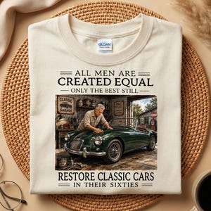Puede incluir: Una camiseta de color crema con un gráfico de una persona puliendo un coche descapotable clásico de color verde oscuro en un garaje. El texto dice: ALL MEN ARE CREATED EQUAL ONLY THE BEST STILL RESTORE CLASSIC CARS IN THEIR SIXTIES. Cuello redondo.